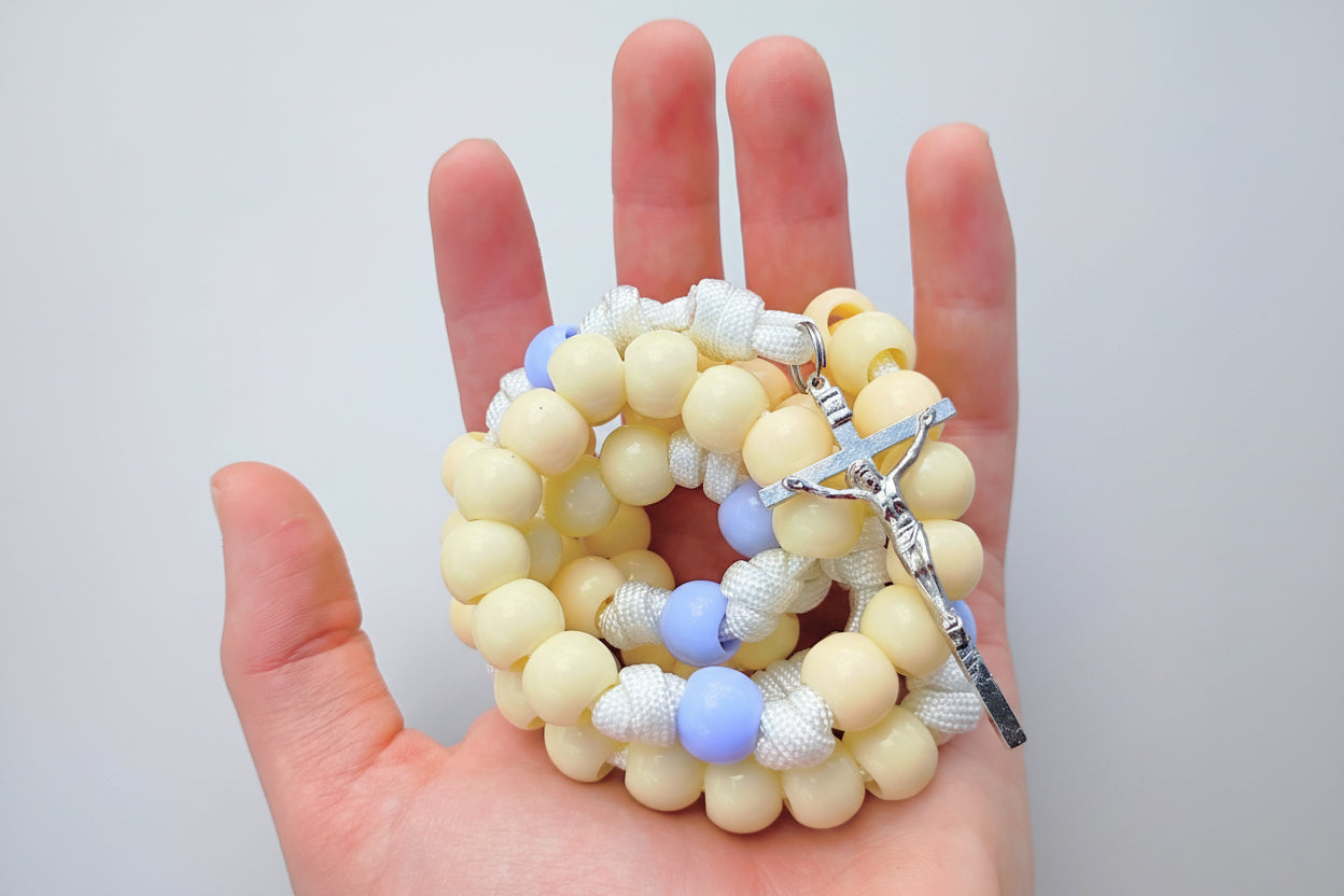 Hand holding a rosary with yellow and blue pastel colored beads on a gray background.