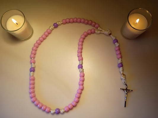 Pink and purple rosary in between two lit candles on a beige surface